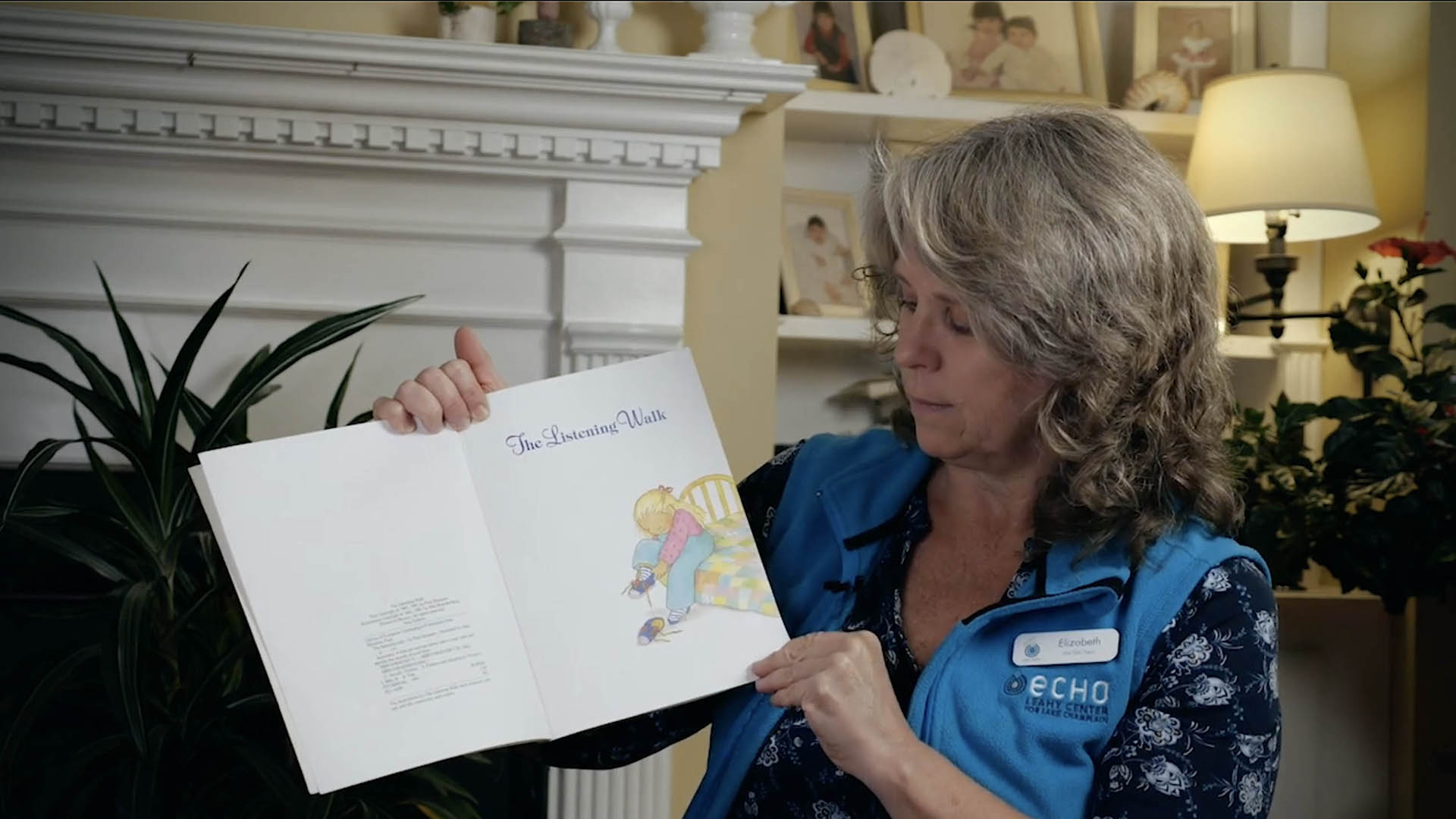 Elizabeth holding a book and about to read. White mantle and lamp in background.