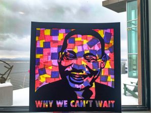 Black laser cut image of MLK with the words "Why We Can't Wait" and behind it a collage of colored paper, hung up on a window with Lake Champlain in the distance