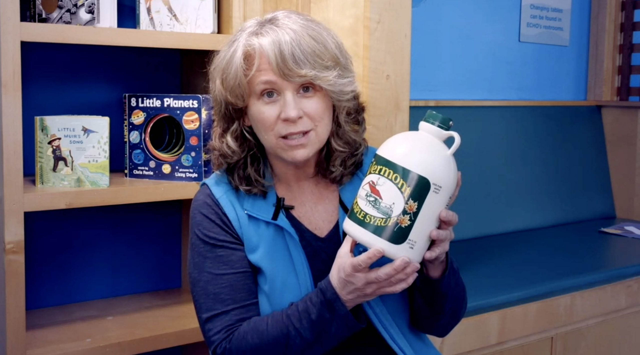 Elizabeth with blonde hair and in an ECHO blue vest holds up a very big jar of maple syrup.