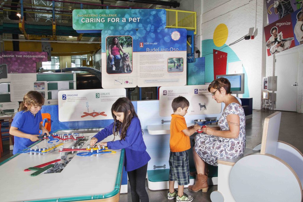 Family with older female adult and three younger kids are at tables in the Human Plus exhibit and are caring for a pet.