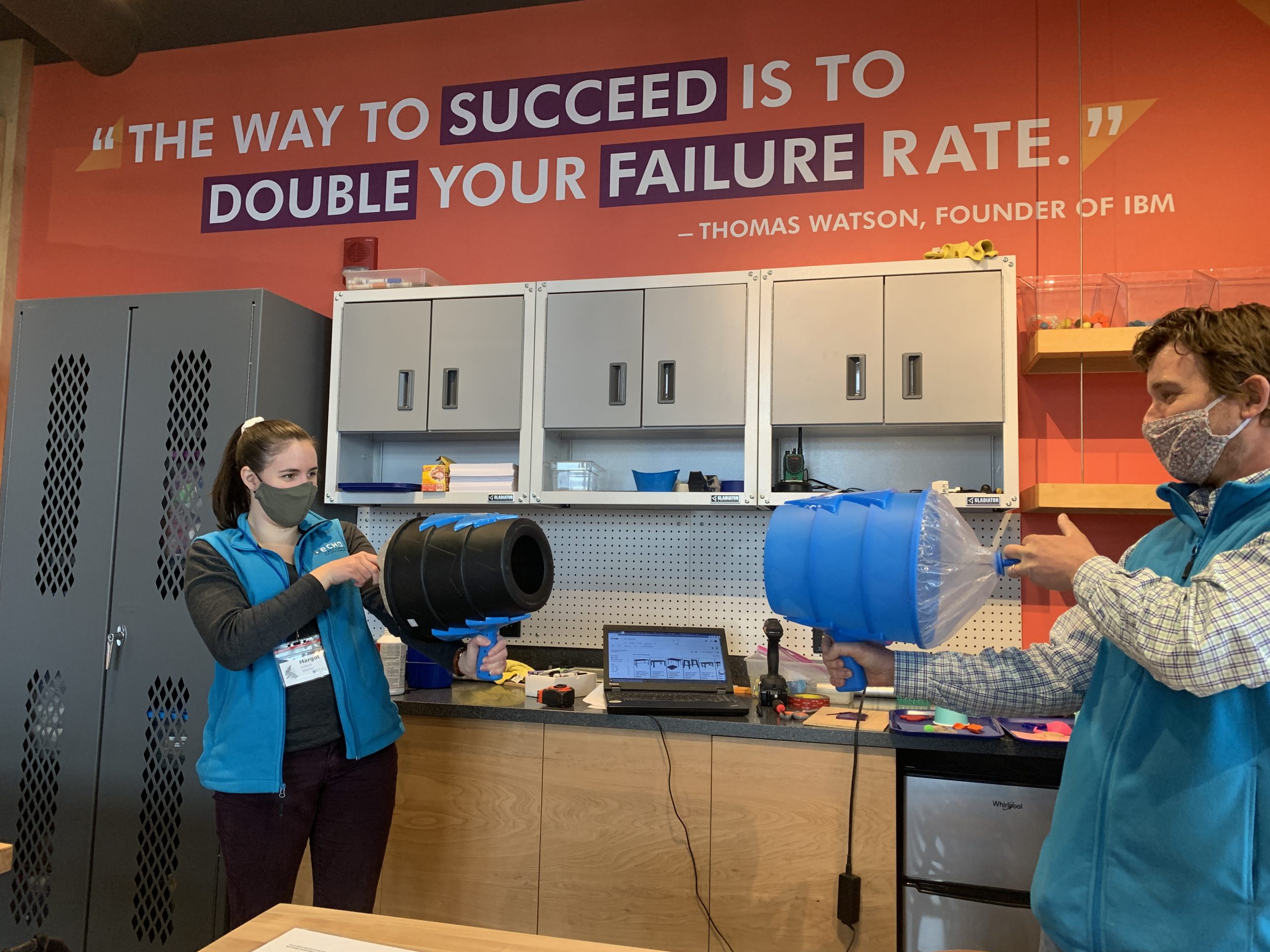 One female ECHO intern in a blue vest aiming an air cannon at a male ECHO educator in a blue vest with an air cannon ready to blow air at the intern. Both people are in front of an orange wall and white cabinets in the Engineer It space.