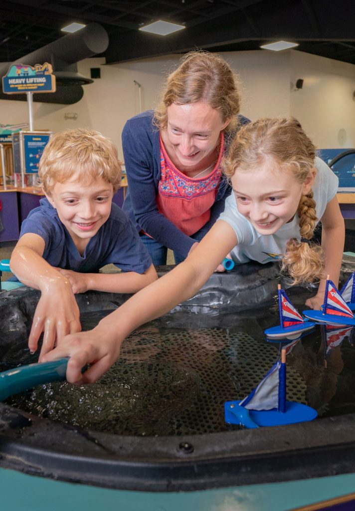 Mom and 2 children are smiling while playing with an exhibit interactive with water and boats.