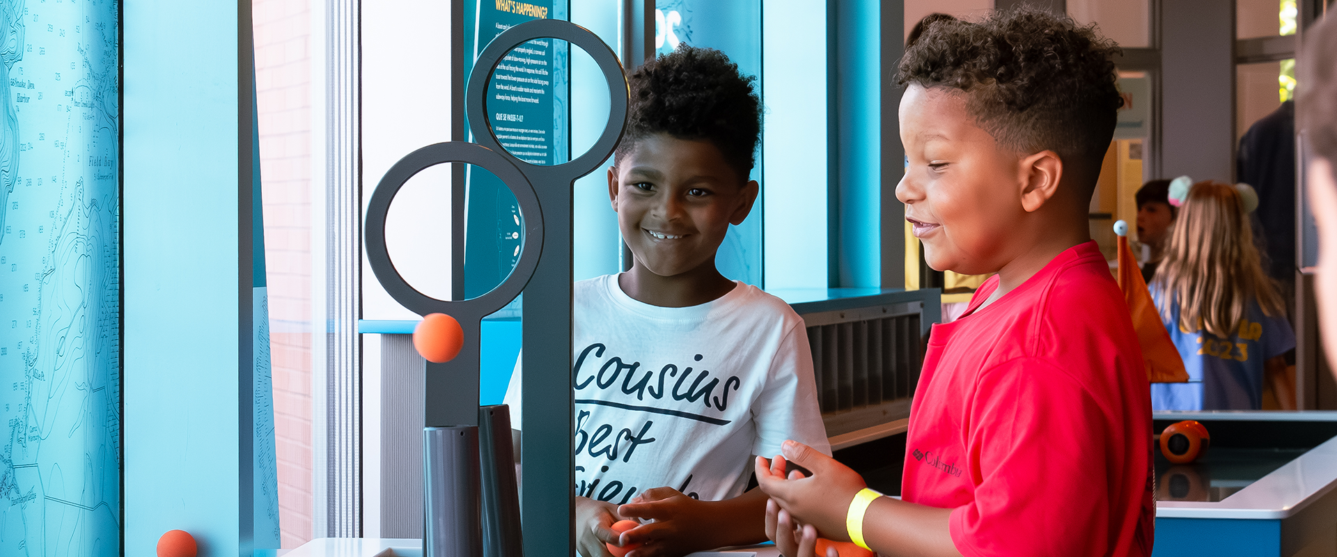 Two young boys use an air tube in an exhibit to hover an orange ball in the air.