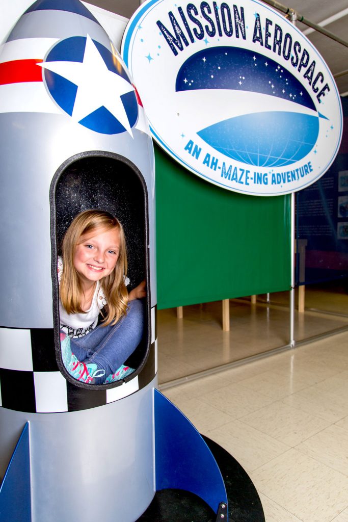 A young girl smiles from inside a model rocket at Mission Aerospace