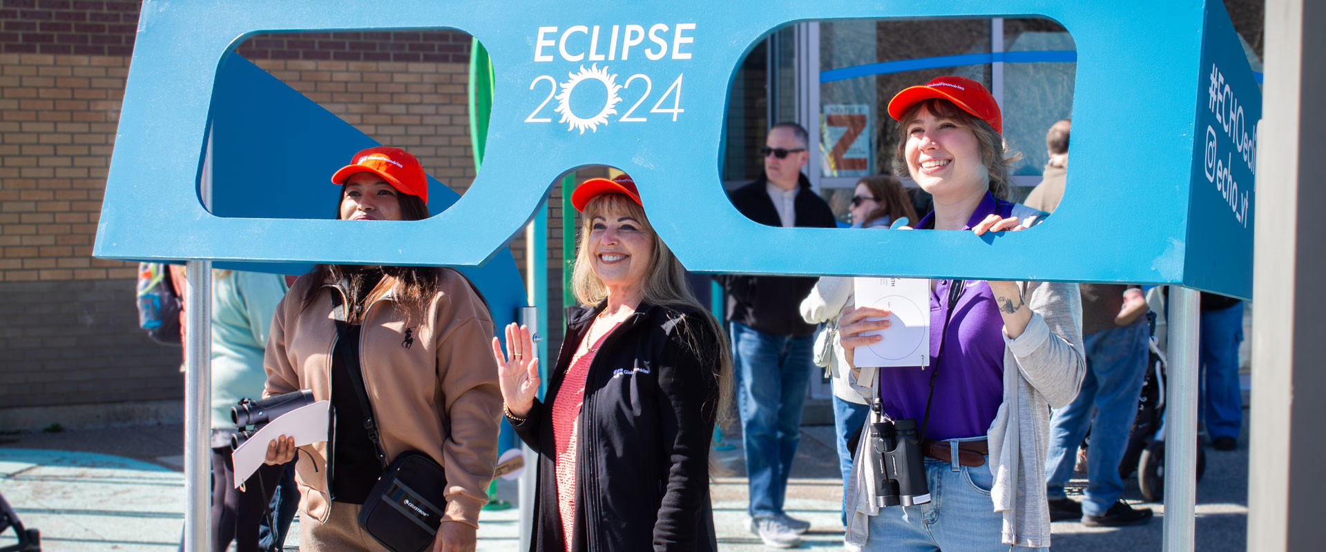 Three Global Foundries volunteers pose with a pair of giant eclipse glasses outside of ECHO