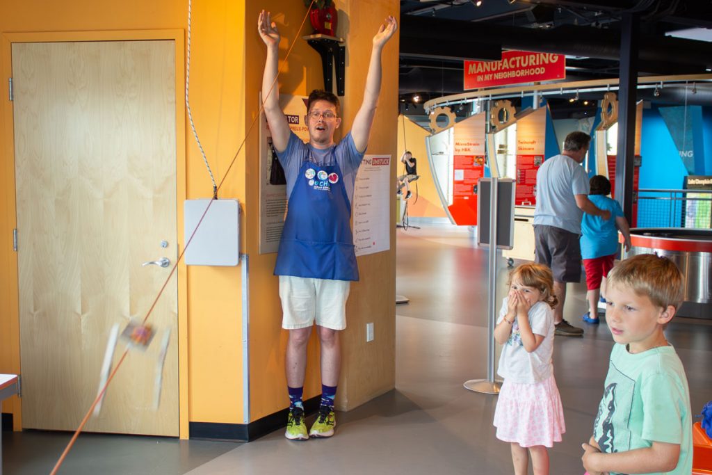 ECHO Educator Billy cheers with their hands in the air as a small handmade zip cart flies down a zipline in Engineer It. Two young children are watching with excitement.