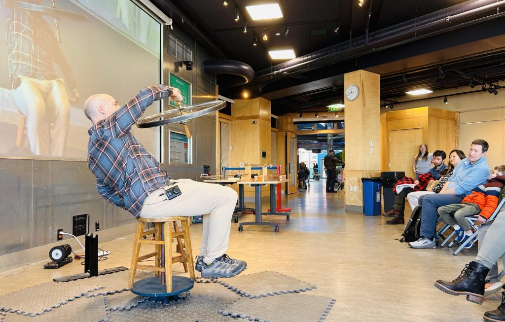 Professor sitting on a stool while spinning and holding a bike wheel while doing a physics demo at ECHO in front of visitors.