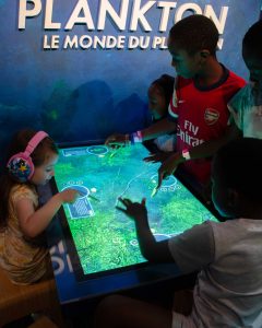 Visitors of all ages sit around the World of Plankton tabletop interactive screen.